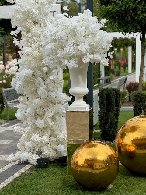 Elegancka dekoracja plenerowa w biało-złotej stylistyce, idealna na luksusowe wesele lub przyjęcie. Na pierwszym planie duża, biała waza na złotym postumencie, wypełniona bujną kompozycją z białych hortensji i orchidei. Obok znajduje się wysoka, kwiatowa aranżacja w formie kolumny, opleciona gęstymi białymi kwiatami. Na trawniku w hotelu Rubin Radom leżą duże, złote, lustrzane kule, dodające nowoczesnego i ekskluzywnego charakteru. W tle widać starannie przycięte krzewy, pergole i alejki, tworzące romantyczną atmosferę w otoczeniu zieleni
