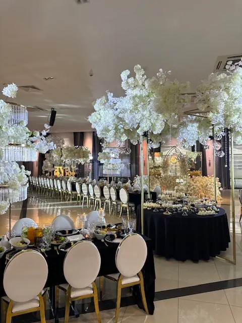 Elegancka sala bankietowa w hotelu Rubin Radom w luksusowym stylu biało-złotym. Długie stoły z czarnymi obrusami są udekorowane białymi kwiatami, kryształowymi świecznikami i złotymi akcentami. Nad stołami wiszą spektakularne kompozycje z białych hortensji i orchidei, dodając wnętrzu romantycznego charakteru. Krzesła tapicerowane białą skórą ze złotymi nogami podkreślają ekskluzywny styl. W tle widać bogato zastawiony stół z eleganckimi deserami oraz złotą dekoracją. Całość tworzy wytworną atmosferę idealną na wesele lub wyjątkową uroczystoś