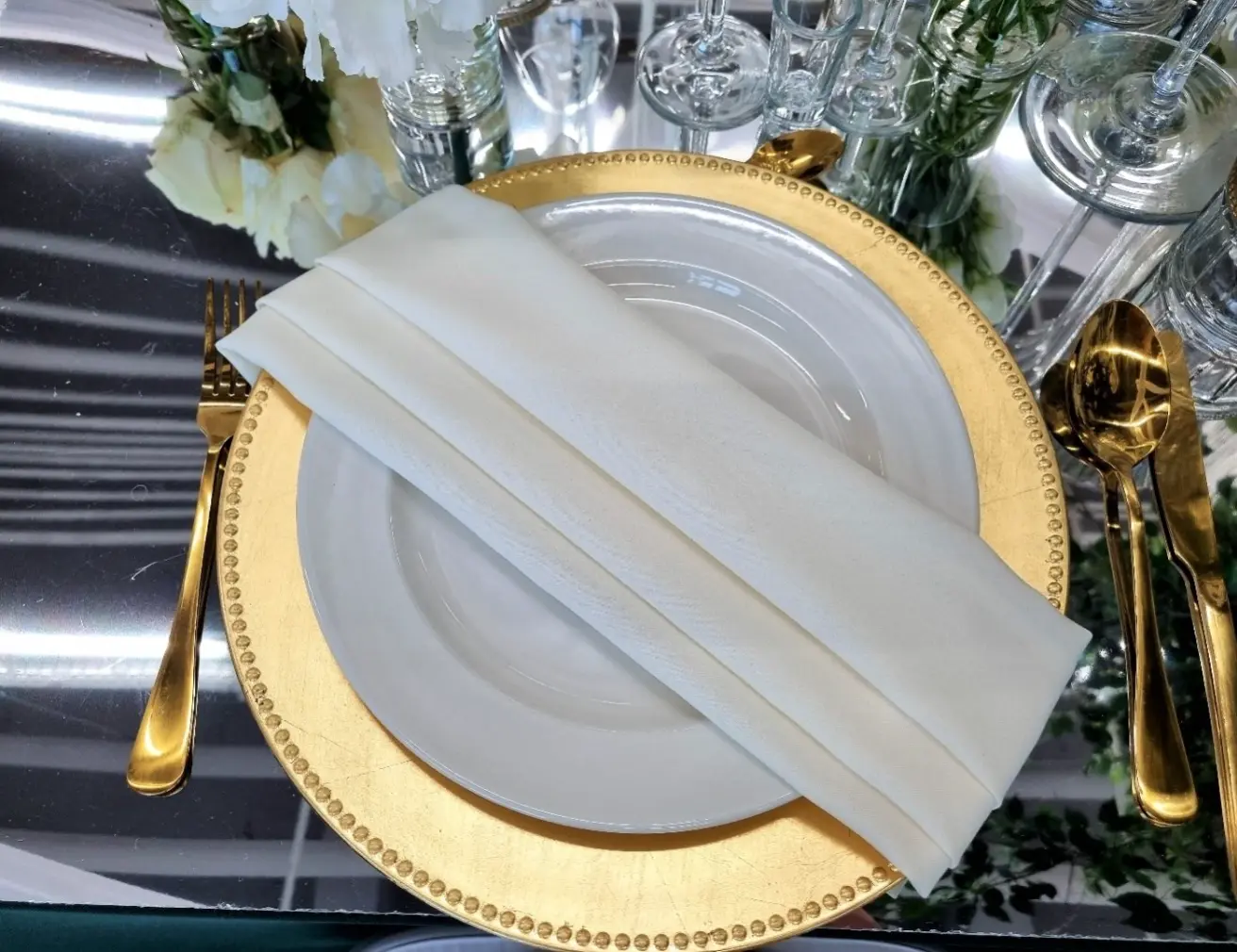 Eleganckie nakrycie stołu z białym talerzem na złotej podkładce z koralikowym brzegiem. Na talerzu znajduje się starannie złożona biała serwetka. Po lewej stronie talerza leży złoty widelec, a po prawej złota łyżka i nóż. W tle widoczna jest elegancka dekoracja kwiatowa oraz kieliszki, co tworzy luksusową atmosferę. Całość prezentuje się subtelnie i elegancko, idealna na wyjątkowe okazje
