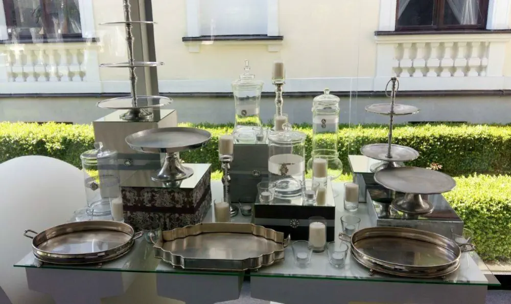 Zestaw eleganckich akcesoriów do serwowania i dekoracji stołu, ustawiony na szklanym blacie. W skład kompozycji wchodzą srebrne patery na torty i ciasta, metalowe tace w różnych kształtach, wysokie słoje aptekarskie z pokrywkami, świeczniki oraz dekoracyjne świece. Idealna aranżacja do candy baru lub stołu słodkości na wesele, bankiet lub przyjęcie tematyczne w stylu glamour, vintage lub klasycznym