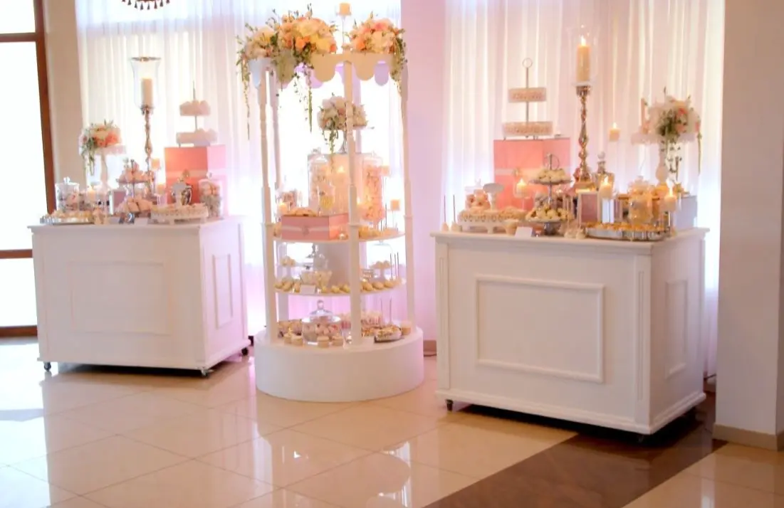 Biała, elegancka aranżacja candy baru na wesele lub przyjęcie, składająca się z trzech stacji – dwóch prostokątnych bufetów i jednej centralnej, okrągłej, dekorowanej kwiatami w pastelowych odcieniach różu i bieli. Na blatach ustawiono szklane pojemniki z kolorowymi słodyczami, stojaki z babeczkami, ciasteczkami i lizakami na patyczkach, a także świeczniki ze świecami dla stworzenia nastrojowego światła. Tło stanowią białe firany i różowe podświetlenie, nadające romantyczny i luksusowy charakter