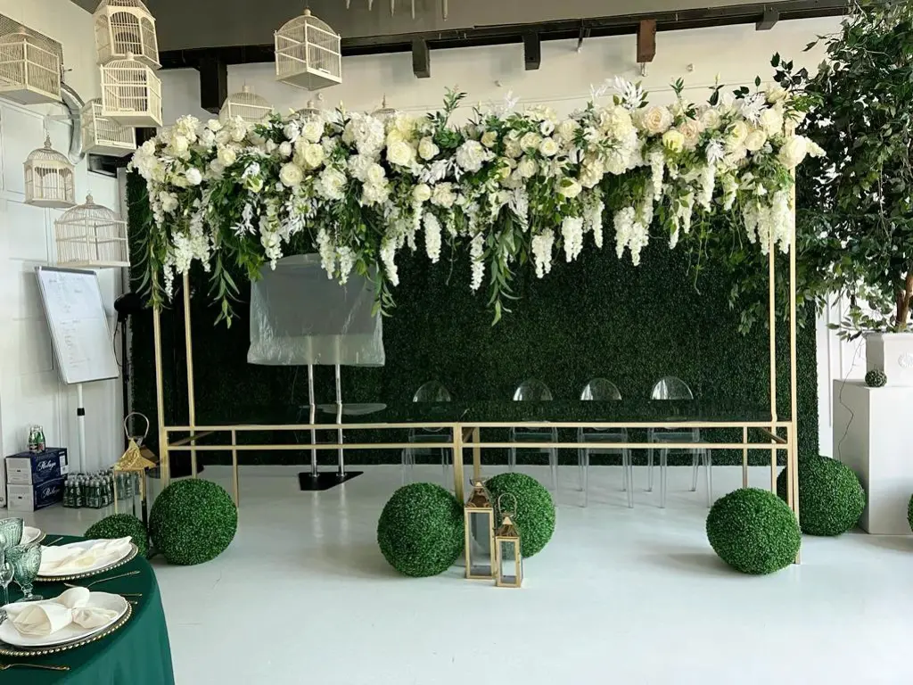 Elegancka dekoracja z kwiatami na specjalną okazję, z białymi różami, liliami i delikatnym zielonym liściem, zawieszona na złotej ramie, tworząc efektowną girlandę. Tło stanowi bujna zielona ściana z sztucznych roślin, która podkreśla naturalny wygląd aranżacji. Przed sceną znajdują się zielone, okrągłe kule z roślinami przypominającymi bukszpan oraz ozdobne lampiony w złotych ramach. Dodatkowo widoczne są lekkie przezroczyste krzesła i eleganckie stoły nakryte białymi serwetkami i zielonymi obrusami, które dodają całości nowoczesnego, a zarazem eleganckiego charakteru