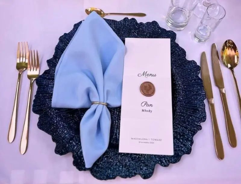 Na eleganckim stole znajduje się talerz o wyraźnym, granatowym kolorze z dekoracyjnym brzegiem, na którym spoczywa błękitna serwetka z ozdobnym pierścieniem, a obok leży menu, którego tytuł MENU jest napisany ozdobnym fontem, a poniżej znajduje się imię Pana Młodego. Całość dopełniają złote sztućce i kieliszki, tworząc wyjątkową atmosferę na uroczystości. Zestaw dobrze współgra z subtelnymi detalami, takimi jak kolorystyka i akcenty w postaci symbolu