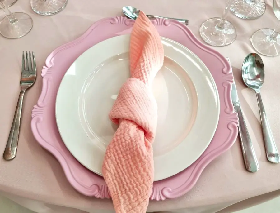 Na stole znajduje się elegancko przygotowane nakrycie. W centrum znajduje się talerz na różowym, ozdobnym talerzu. Na talerzu leży różowa serwetka, starannie złożona i owinięta wokół talerza. Po lewej stronie talerza znajduje się widelec, a po prawej łyżka. W tle widoczna jest kieliszek do wina i inne akcesoria stołowe. Stół pokryty jest jasnoróżowym obrusem, a całość tworzy elegancką, stylową aranżację stołową