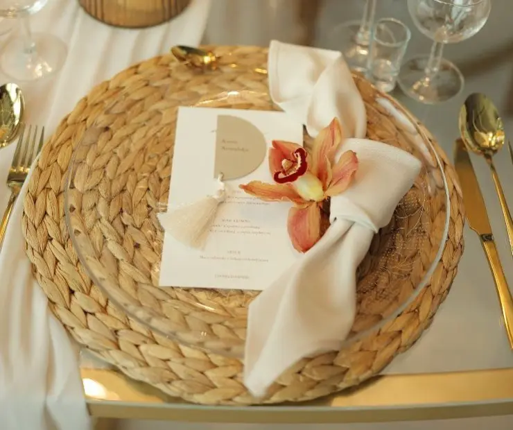Na stole znajduje się elegancko nakryte miejsce. W centrum widoczny jest przezroczysty talerz na okrągłej, plecionej podkładce z naturalnego materiału. Na talerzu leży elegancko złożona biała serwetka, ozdobiona kwiatem orchidei w odcieniach pomarańczowego i czerwonego, oraz małą, białą frędzelkową ozdobą. Obok serwetki znajduje się karta z menu, umieszczona na talerzu. Po prawej stronie talerza widoczna jest złota łyżka, a obok kieliszki do wina