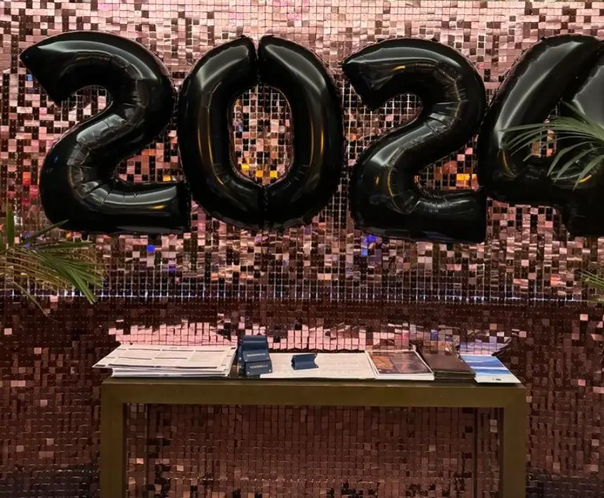 Efektowna dekoracja noworoczna lub eventowa z połyskującym, różowo-złotym tłem z paneli lustrzanych typu shimmer wall. Na środku zawieszono duże, czarne balony foliowe tworzące napis „2024”. Przed ścianką ustawiono prosty, drewniany stolik z dokumentami, katalogami i materiałami promocyjnymi, sugerujący funkcję recepcyjną lub informacyjną. Dekoracja idealna na sylwestra, galę firmową, konferencję lub imprezę powitalną w eleganckim, nowoczesnym stylu