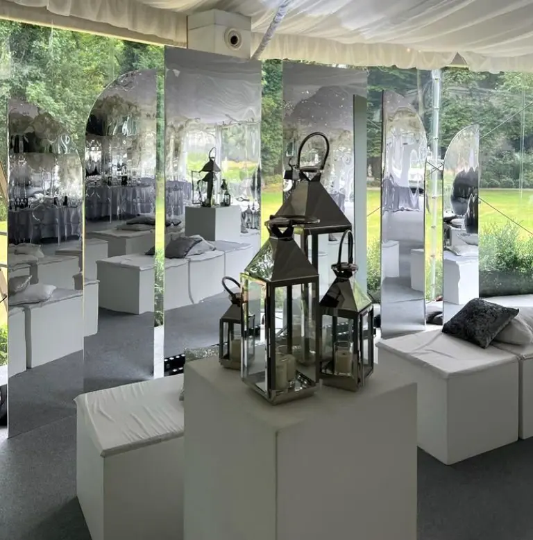 Elegancka strefa chillout na przyjęciu plenerowym, urządzona w nowoczesnym stylu glamour. Na pierwszym planie białe siedziska i podesty z dekoracyjnymi latarniami ze szkła i metalu, w których umieszczono świece LED. Tło stanowią wysokie, lustrzane panele o prostokątnym i łukowym kształcie, które odbijają otoczenie i dodają przestrzeni głębi. Całość znajduje się pod białym namiotem z materiałowym sufitem, tworząc luksusową atmosferę idealną na wesele, bankiet lub garden party