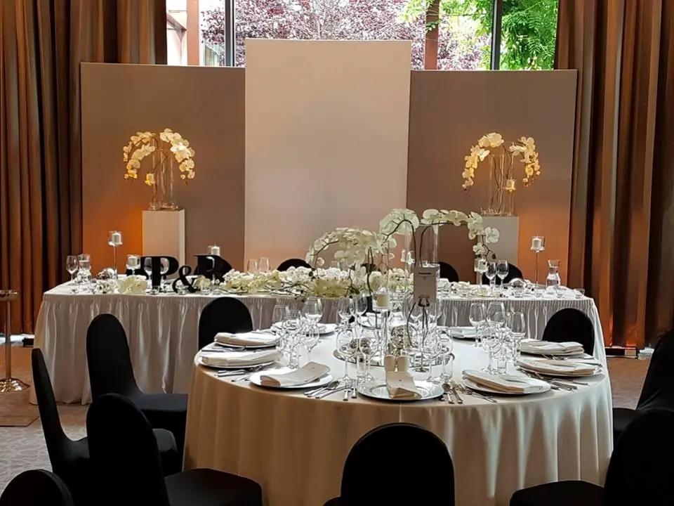 Nowoczesna dekoracja sali weselnej w eleganckim stylu minimalistycznym. Centralne miejsce zajmuje stół pary młodej z białym obrusem, ozdobiony białymi storczykami i inicjałami P & R w czerni. Za nim znajduje się biały panel ślubny w towarzystwie dwóch podświetlanych kolumn z eleganckimi kompozycjami z orchidei. Przed nim ustawiony okrągły stół gościnny z czarnymi krzesłami, srebrną zastawą i nowoczesnymi dekoracjami. Całość utrzymana w biało-czarnej kolorystyce z akcentami glamour