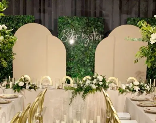 Elegancka dekoracja weselna z napisem „Better Together” na tle paneli z zielonego bukszpanu i kremowych ścianek w zaokrąglonym kształcie. Przestrzeń ozdobiona białymi i zielonymi kompozycjami kwiatowymi, złotymi krzesłami z białymi poduszkami i świecami w szklanych osłonach. Styl glamour z elementami natury, idealny na wesele lub uroczystą kolację