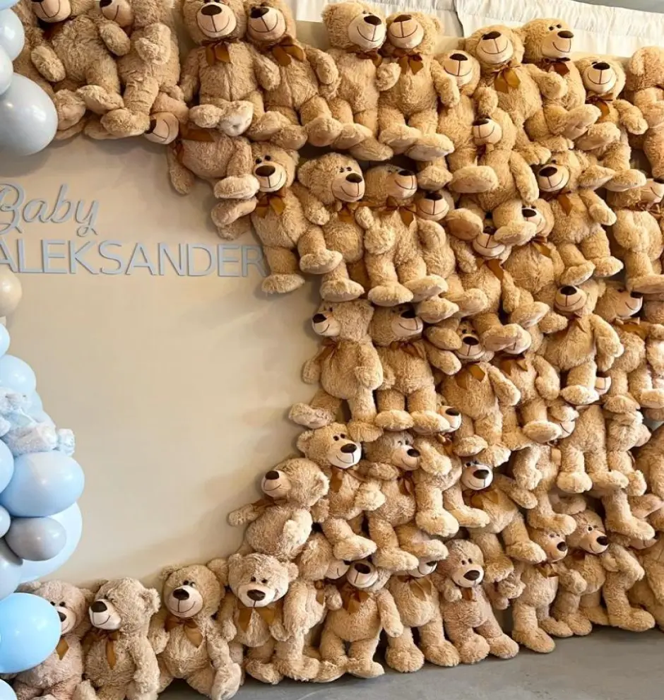 Urocza dekoracja na baby shower lub przyjęcie powitalne dla dziecka, ozdobiona dziesiątkami pluszowych misiów w kolorze jasnobrązowym z brązowymi kokardkami. Maskotki tworzą efektowną ramę wokół jasnego tła z napisem Baby Aleksander. Po lewej stronie znajduje się delikatna girlanda z pastelowych balonów w odcieniach błękitu i bieli. Całość tworzy ciepłą, przytulną atmosferę idealną na imprezę dla malucha