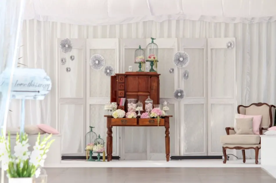 Vintage'owa dekoracja ślubna z romantycznym akcentem w stylu shabby chic. W centrum ustawiono drewniane biurko z zabytkową nadstawką, udekorowane słoikami wypełnionymi różowymi i białymi kwiatami, cukierkami oraz świecznikami. Na blacie i obok mebla znajdują się metalowe klatki z dodatkowymi kwiatami. Tło stanowią białe, stylizowane drzwi ustawione w rzędzie, udekorowane delikatnymi papierowymi rozetami w odcieniach szarości. Całość uzupełnia kremowy fotel z różowymi poduszkami oraz biały baldachim zawieszony nad dekoracją, nadający aranżacji miękkości i romantyzmu. Dekoracja idealna na ślub w stylu rustykalnym lub boho