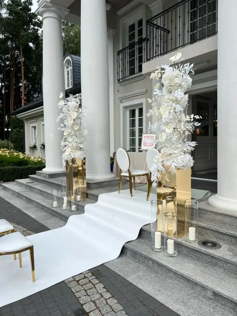 Luksusowa aranżacja ślubna na zewnątrz eleganckiego budynku z kolumnami, z centralnie umieszczoną białą ścieżką prowadzącą na schody. Po obu stronach stoją wysokie, złote postumenty z artystycznymi kompozycjami z białych sztucznych kwiatów, liści i piór. Obok znajdują się transparentne tuby z dużymi białymi świecami. Ustawione są także białe krzesła z pozłacanymi nogami i oparciami.