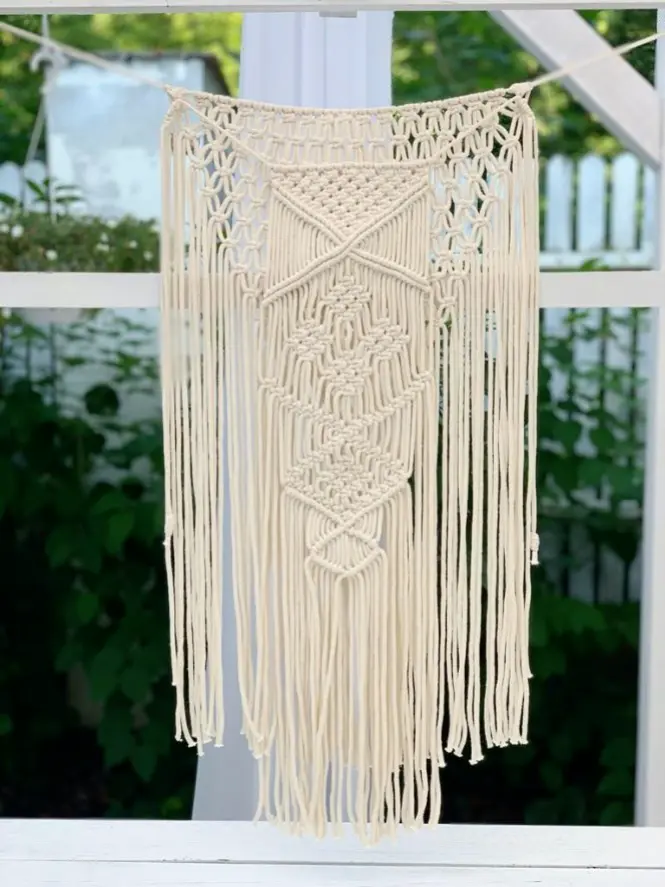 Ręcznie pleciona makrama z jasnego sznurka bawełnianego w stylu boho, zawieszona na białej konstrukcji na tle zieleni ogrodu. Wzór składa się z geometrycznych splotów, rombów i węzłów, a dolna część zakończona jest długimi luźnymi frędzlami. Dekoracja idealna do wnętrz w stylu naturalnym, rustykalnym, a także jako ozdoba na wesele, taras lub sesję zdjęciową w plenerze