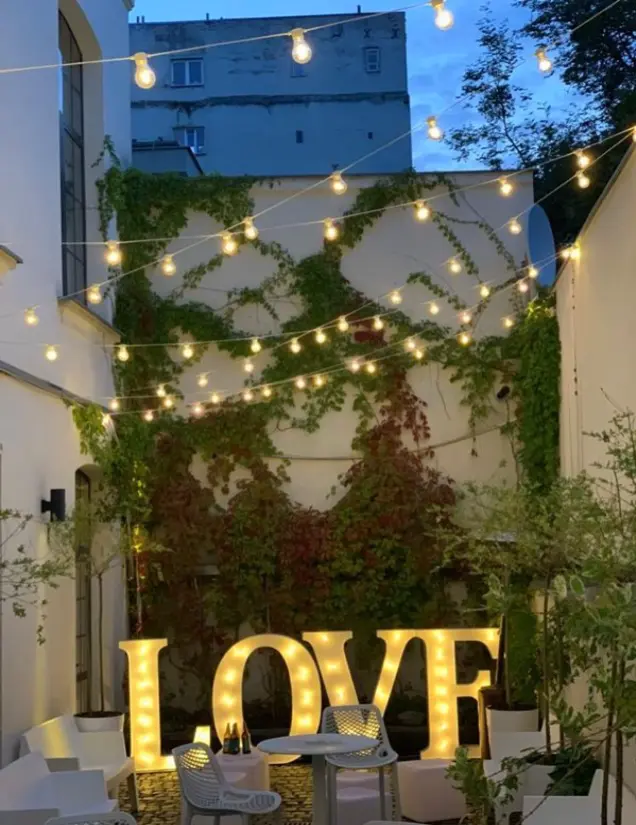 Romantyczna aranżacja dziedzińca w stylu boho glamour z podświetlanym napisem „LOVE” w dużym formacie, ustawionym na tle porośniętej bluszczem ściany. Nad przestrzenią rozciągnięte są girlandy z żarówek dających ciepłe światło, tworzące nastrojowy klimat. Na dziedzińcu znajdują się białe meble ogrodowe, w tym stolik i krzesła, a całość otaczają ozdobne rośliny w donicach. Idealne miejsce na romantyczną kolację, wesele, zaręczyny lub sesję zdjęciową o zachodzie słońca