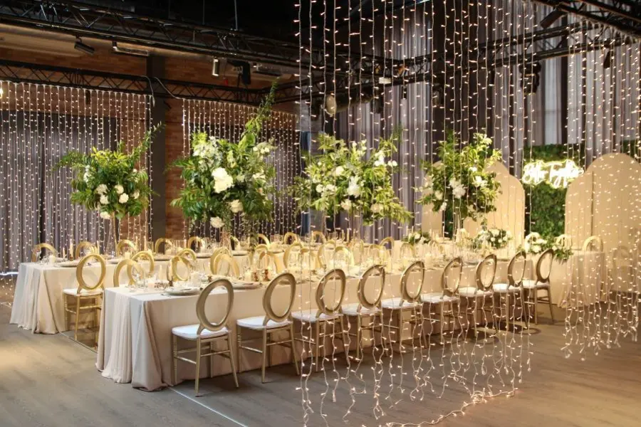 Elegancka sala weselna udekorowana w stylu glamour z nutą romantyzmu. Stoły w kształcie litery U nakryte kremowymi obrusami, ozdobione są wysokimi kompozycjami z zieleni i białych kwiatów. Złote krzesła z białymi siedziskami podkreślają luksusowy charakter aranżacji. Całość otoczona jest kurtynami świetlnymi z drobnych lampek LED, tworzącymi magiczny klimat. W tle ścianka z zieleni z neonem „Better Together” i pastelowe panele. Idealna dekoracja na wesele, przyjęcie lub uroczystą kolację w wyrafinowanym stylu
