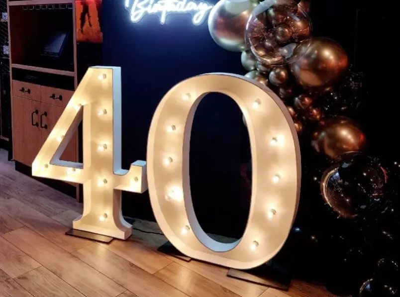 Efektowna dekoracja urodzinowa z dużymi, podświetlanymi cyframi „40”, symbolizującymi czterdzieste urodziny. Jasne litery w stylu retro ozdobione są żarówkami i ustawione na drewnianej podłodze w eleganckim wnętrzu. W tle widoczna czarna ściana z napisem neonowym „Birthday” oraz aranżacją z metalicznych balonów w odcieniach złota, miedzi i czerni. Styl glamour z nutą nowoczesności, idealny na luksusową imprezę urodzinową