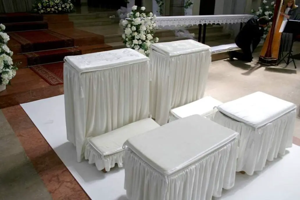 Zestaw ślubnych klęczników i ławek obitych białym materiałem z falbanami, ustawiony w kościele na białym dywanie, przeznaczony dla pary młodej oraz świadków podczas ceremonii zaślubin. Tło udekorowane jest białymi kwiatami i klasycznymi dywanami prowadzącymi do ołtarza, co podkreśla elegancki, sakralny charakter uroczystości.