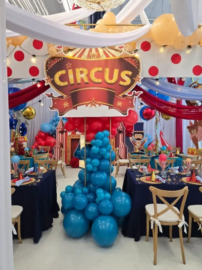 Kolorowa dekoracja wnętrza na tematyczne przyjęcie w stylu cyrkowym. Centralnie zawieszony duży szyld z napisem „CIRCUS” w stylu retro, pod nim kolumna z niebieskich balonów. W tle makieta namiotu cyrkowego, postacie animatorów i dekoracje balonowe w czerwieni, niebieskim i złocie. Stoły udekorowane granatowymi obrusami, kolorowymi czapkami imprezowymi i akcentami w stylu cyrkowym. Nad sufitem białe i czerwone materiały oraz żółte balony z girlandą świetlną, tworzące dynamiczne, festynowe zadaszenie. Całość oddaje radosny klimat dziecięcej imprezy urodzinowej z motywem cyrku