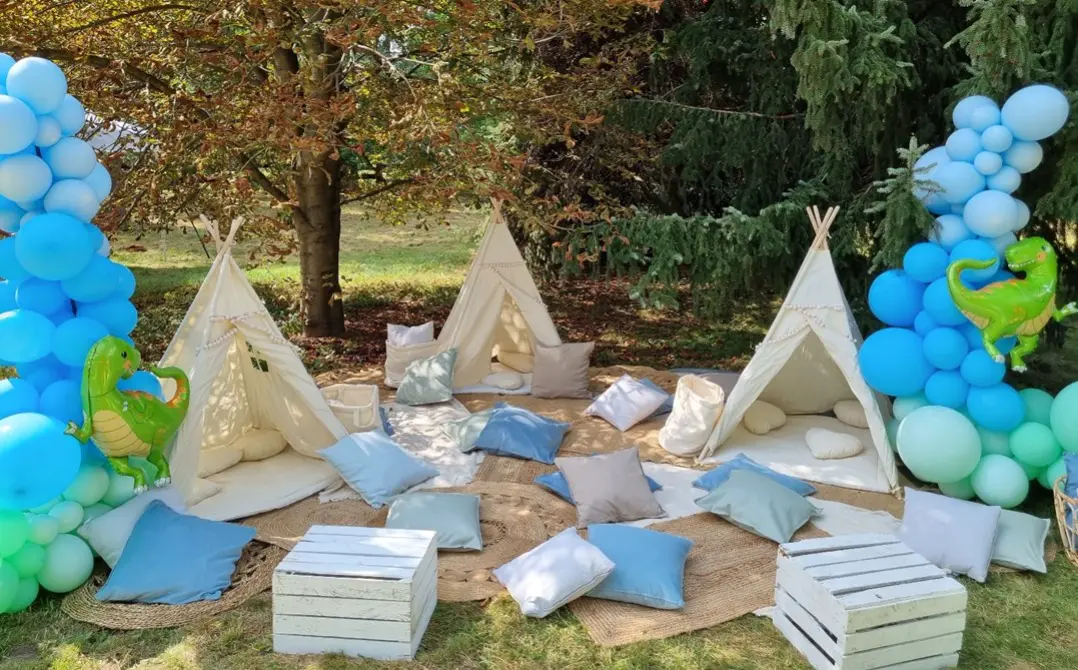 Dekoracyjna strefa relaksu na świeżym powietrzu w stylu tipi, z trzema białymi namiotami tipi ustawionymi na trawie, otoczonymi wiklinowymi matami, jasnymi i niebieskimi poduszkami oraz białymi drewnianymi skrzynkami pełniącymi funkcję stolików. Po bokach znajdują się duże kolumny z niebieskimi i miętowymi balonami oraz zielone dmuchane dinozaury jako ozdoby. Całość usytuowana w cieniu drzew, tworzy przyjazną i komfortową przestrzeń idealną na dziecięce przyjęcie lub piknik tematyczny