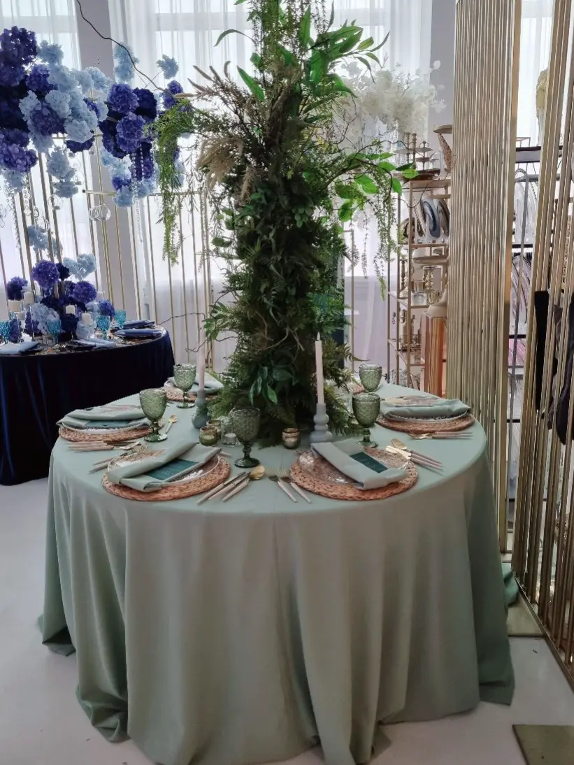 Na zdjęciu widoczna jest wyjątkowa aranżacja stołowa w stylu naturalnym i eleganckim. Stół jest przykryty miękkim, zielonym obrusem, a wokół niego rozmieszczone są talerze na plecionych podkładkach, z zielonymi kieliszkami oraz złotymi sztućcami. Na talerzach leżą elegancko złożone serwetki w odcieniu zieleni, spięte ozdobnymi pierścieniami. W centralnej części stołu znajduje się imponująca dekoracja w postaci wysokiej kompozycji z liści, gałęzi i kwiatów, która tworzy naturalny, leśny klimat. Wokół stołu ustawione są świeczniki z długimi świecami, które dopełniają atmosferę. W tle widoczna jest dekoracja z fioletowych i niebieskich kwiatów, które wprowadzają dodatkowy kolorystyczny akcent do całej aranżacji