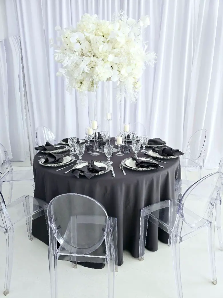 Elegancka aranżacja stołu z wysokim, kwiatowym arrangementem w odcieniach bieli, składającym się z orchidei i innych delikatnych kwiatów. Bukiet umieszczony na szklanej podstawie tworzy wyjątkowy punkt centralny. Stół pokryty jest ciemnoszarym obrusem, a na nim ustawiono biało-czarne talerze, sztućce i kieliszki do wina. Na talerzach znajdują się eleganckie czarne serwetki. Dookoła stołu znajdują się nowoczesne przezroczyste krzesła, które podkreślają minimalistyczny, ale luksusowy charakter aranżacji, a w tle widoczna jest gładka biała tkanina