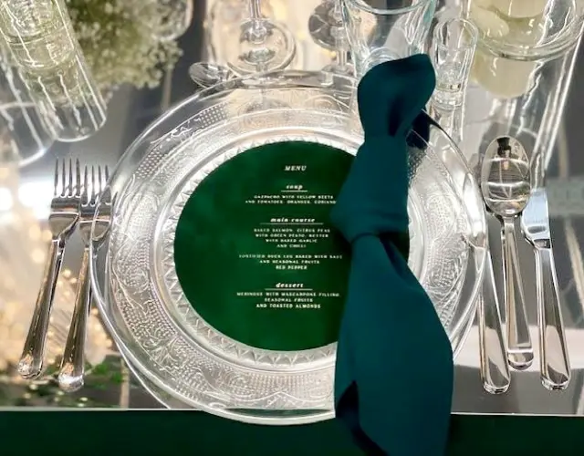 Eleganckie nakrycie stołu z przezroczystym talerzem ozdobionym misternym wzorem. Na talerzu znajduje się ciemnozielona kartka z eleganckim, złotym napisem MENU, zawierająca szczegółowy opis dań: zupy, dania głównego i deseru. Obok talerza znajduje się sztućce – widelec, nóż i łyżka. Na stole leży ciemnozielona serwetka zawiązana w węzeł, a w tle widać kieliszki oraz delikatne dekoracje kwiatowe, tworzące elegancką i stylową atmosferę