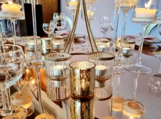Zbliżenie na elegancko udekorowany stół z licznymi świecami w szklanych świecznikach o różnych wysokościach, rozmieszczonymi wokół złotego stojaka. Świece odbijają się w lustrzanej powierzchni blatu, tworząc efektowną grę świateł. W tle widać kieliszki i nakrycia stołowe z ozdobnymi serwetkami, sugerujące przygotowanie do eleganckiego przyjęcia lub kolacji